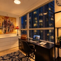 Modern home office in a high-rise apartment: black desk with laptop, notebook, and chair by large windows at dusk, with abstract painting and two lamps behind.