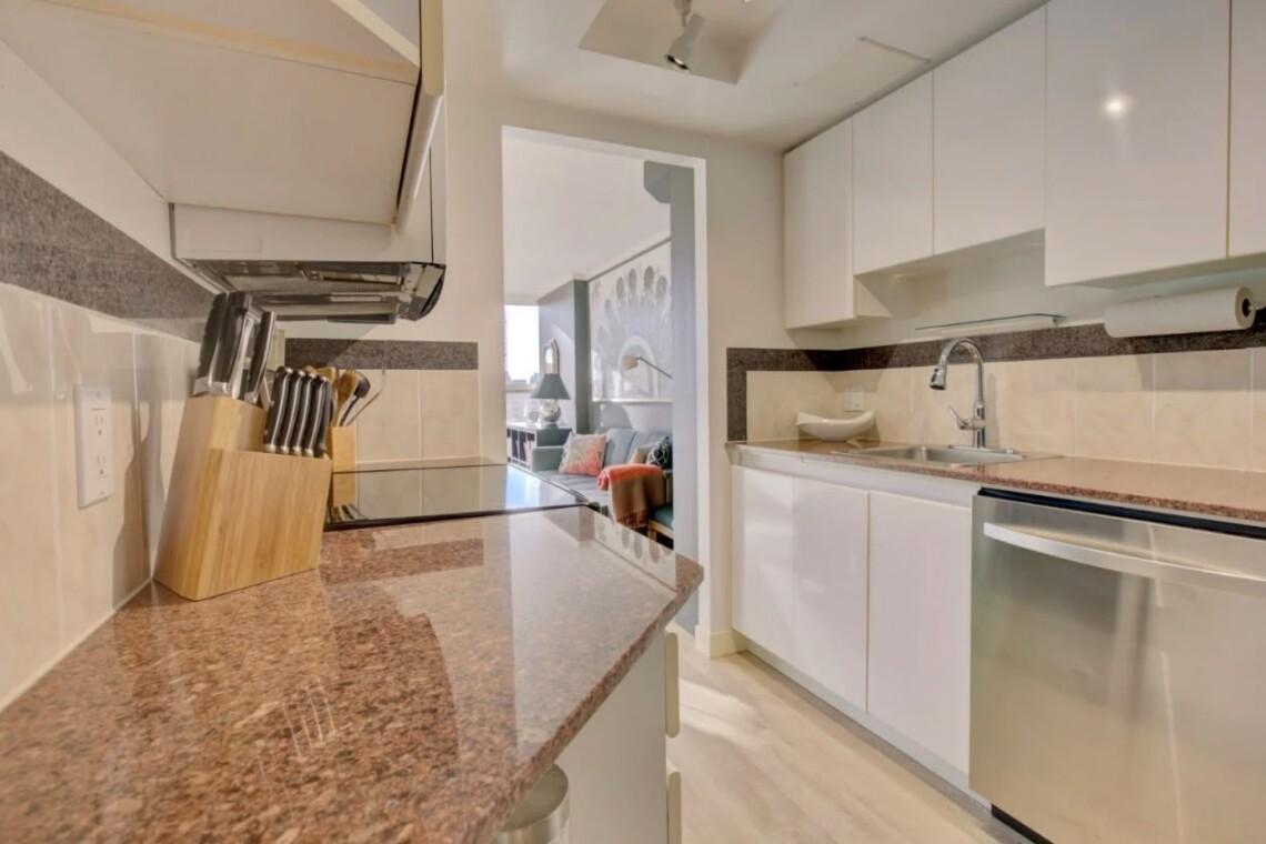 Modern white kitchen with granite countertops, knife block, and a stainless steel dishwasher, opening into a living room.