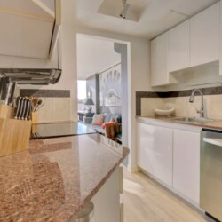 Modern white kitchen with granite countertops, knife block, and a stainless steel dishwasher, opening into a living room.