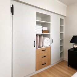 White built-in wardrobe with sliding doors and open middle shelves holding towels and pillows, plus a light wood chest of drawers below.