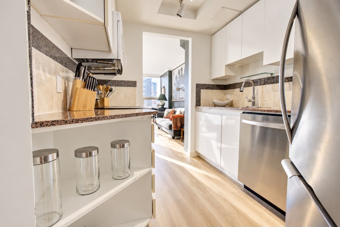 Bright, modern kitchen with white cabinets, granite counters, stainless appliances, and a doorway to a living room.