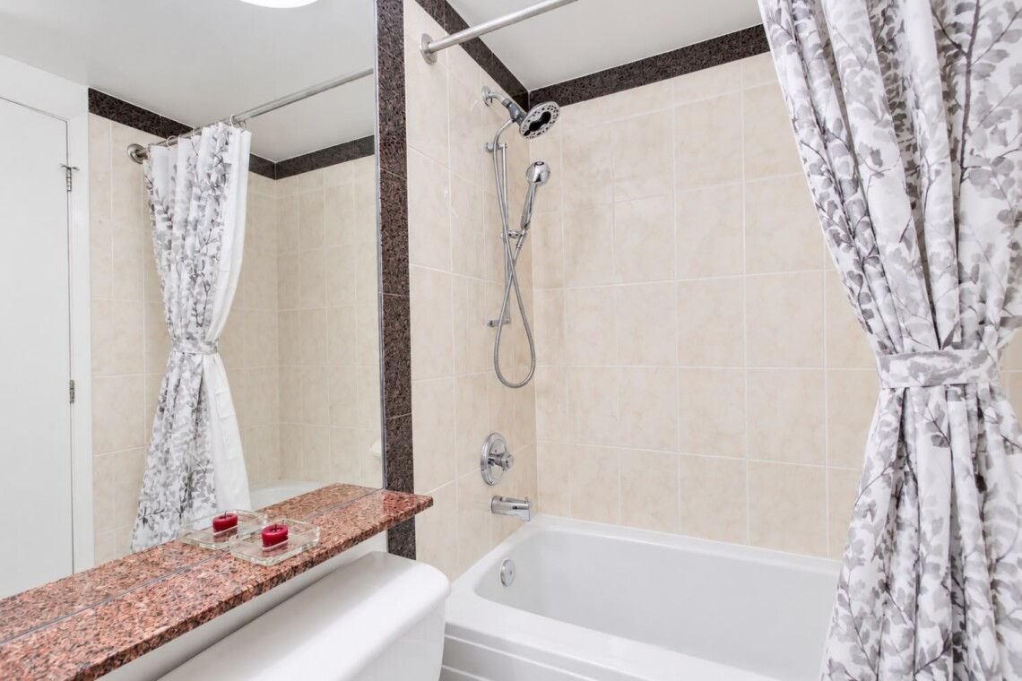 Beige-tiled bathroom with a bathtub, a handheld shower, and gray floral curtains; a granite ledge holds red candles at the edge of the tub.