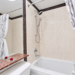 Beige-tiled bathroom with a bathtub, a handheld shower, and gray floral curtains; a granite ledge holds red candles at the edge of the tub.