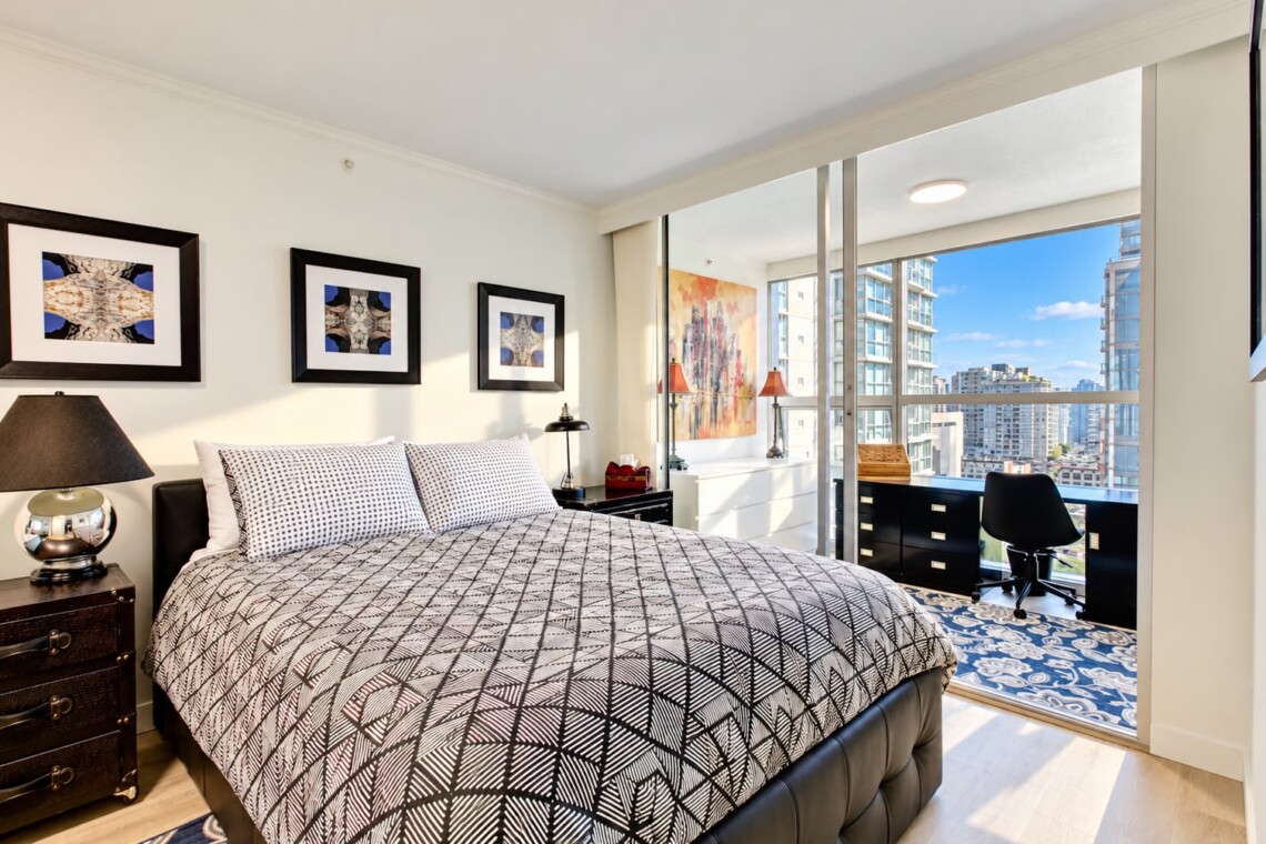 Bright modern bedroom with a geometric-patterned quilt, two nightstands, and framed wall art, opening to a balcony with city views.