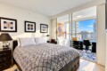 Bright modern bedroom with a geometric-patterned quilt, two nightstands, and framed wall art, opening to a balcony with city views.