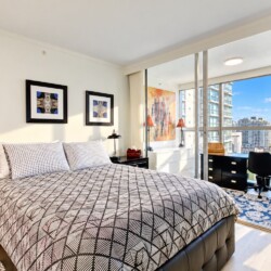 Bright modern bedroom with a geometric-patterned quilt, two nightstands, and framed wall art, opening to a balcony with city views.