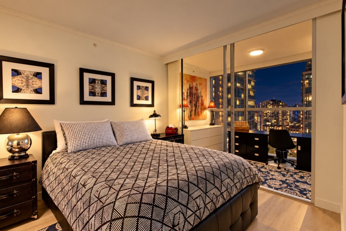 Modern bedroom with geometric duvet, three framed pictures, and a balcony overlooking city lights at night.