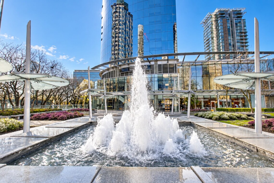 Modern glass office building with a large central fountain and landscaped plaza in front.