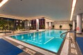 Indoor swimming pool with blue water, no-diving signs along the edge, and blue mats on the tiled floor by a row of chairs and large windows.
