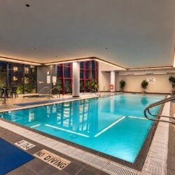 Indoor swimming pool with blue water, no-diving signs along the edge, and blue mats on the tiled floor by a row of chairs and large windows.