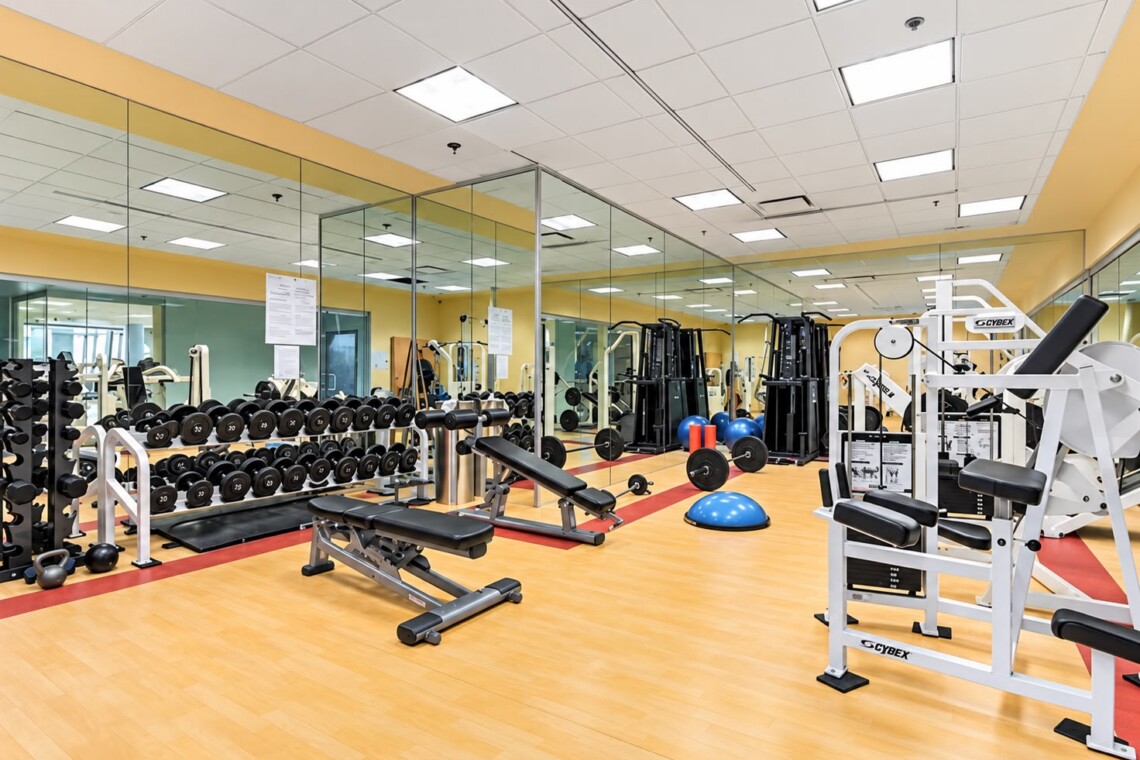 Well-equipped gym with dumbbell racks, benches, and weight machines in a bright, mirrored room with yellow accents.