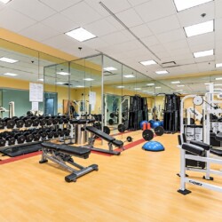 Well-equipped gym with dumbbell racks, benches, and weight machines in a bright, mirrored room with yellow accents.