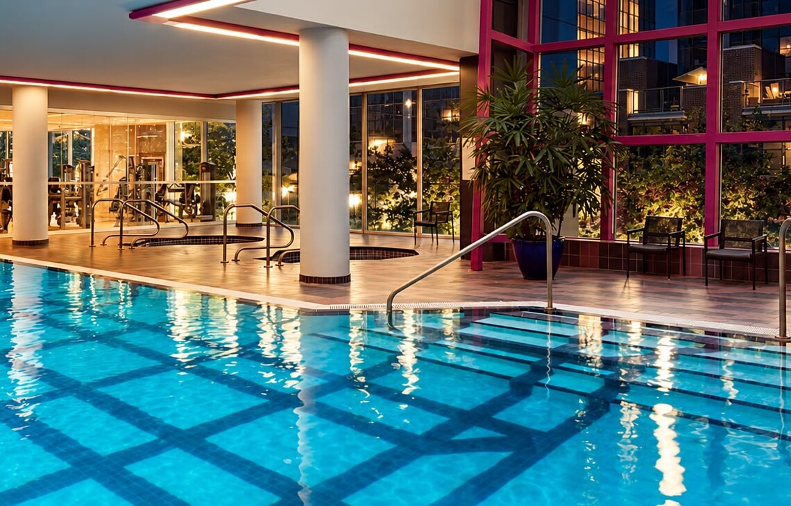 Indoor swimming pool in a fitness center, with the gym visible through glass walls in the background.