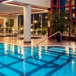 Indoor swimming pool in a fitness center, with the gym visible through glass walls in the background.