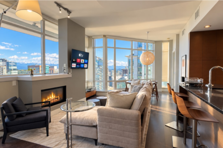 Bright open living room with a beige sofa, glass coffee table, fireplace, and wall-mounted TV; large windows overlook a cityscape.